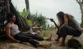 Brad (Dylan O'Brien), a barefoot, bedraggled man in ripped pants and a filthy sleeveless T-shirt, sprawls on sand in front of a shelter made out of fronds and leaves, as Linda (Rachel McAdams), a similarly grimy woman, gestures at him with a knife in Sam Raimi's Send Help