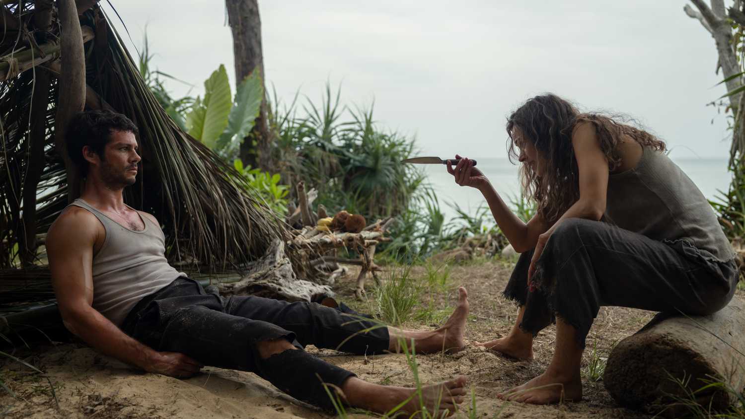 Brad (Dylan O'Brien), a barefoot, bedraggled man in ripped pants and a filthy sleeveless T-shirt, sprawls on sand in front of a shelter made out of fronds and leaves, as Linda (Rachel McAdams), a similarly grimy woman, gestures at him with a knife in Sam Raimi's Send Help