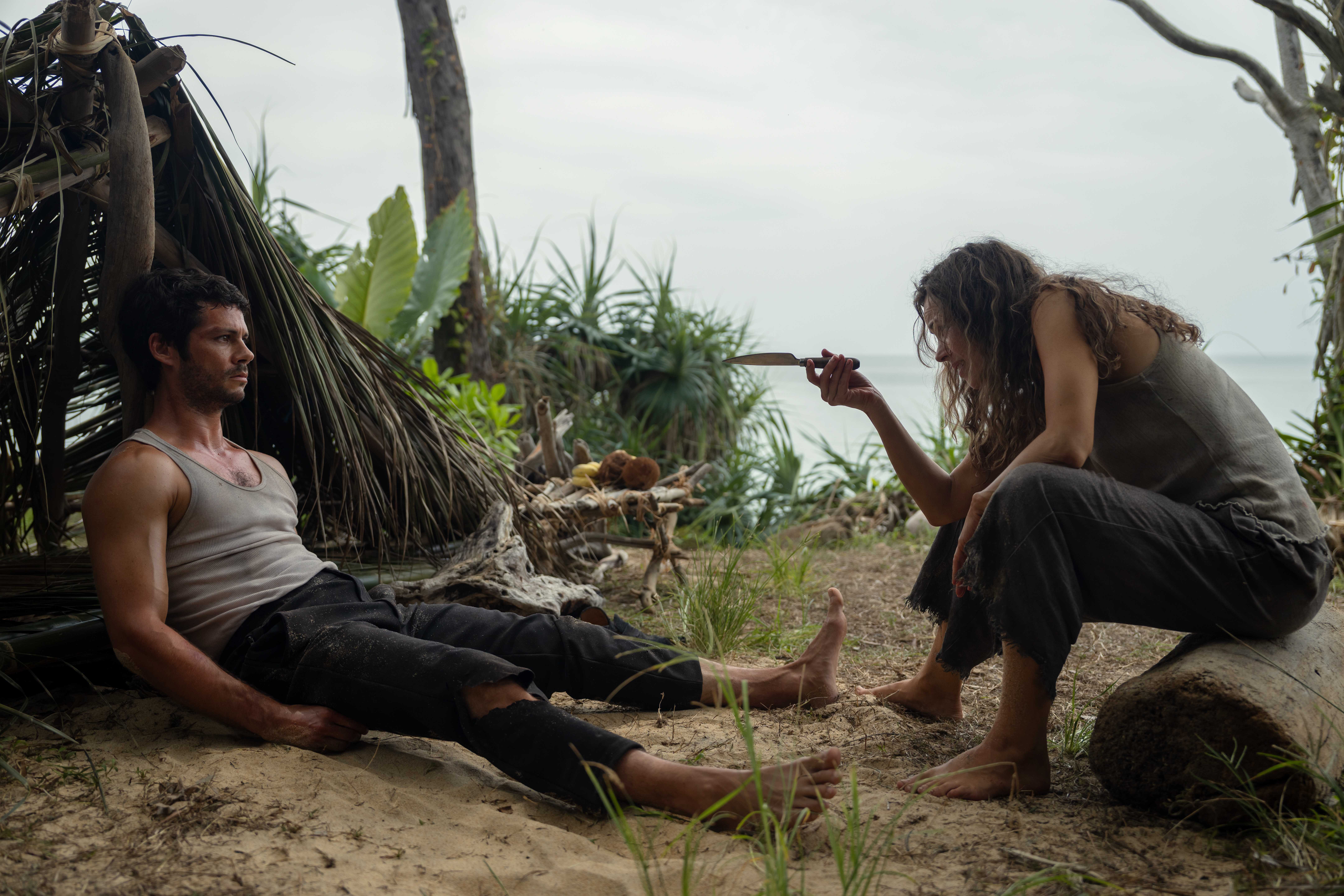 Brad (Dylan O'Brien), a barefoot, bedraggled man in ripped pants and a filthy sleeveless T-shirt, sprawls on sand in front of a shelter made out of fronds and leaves, as Linda (Rachel McAdams), a similarly grimy woman, gestures at him with a knife in Sam Raimi's Send Help