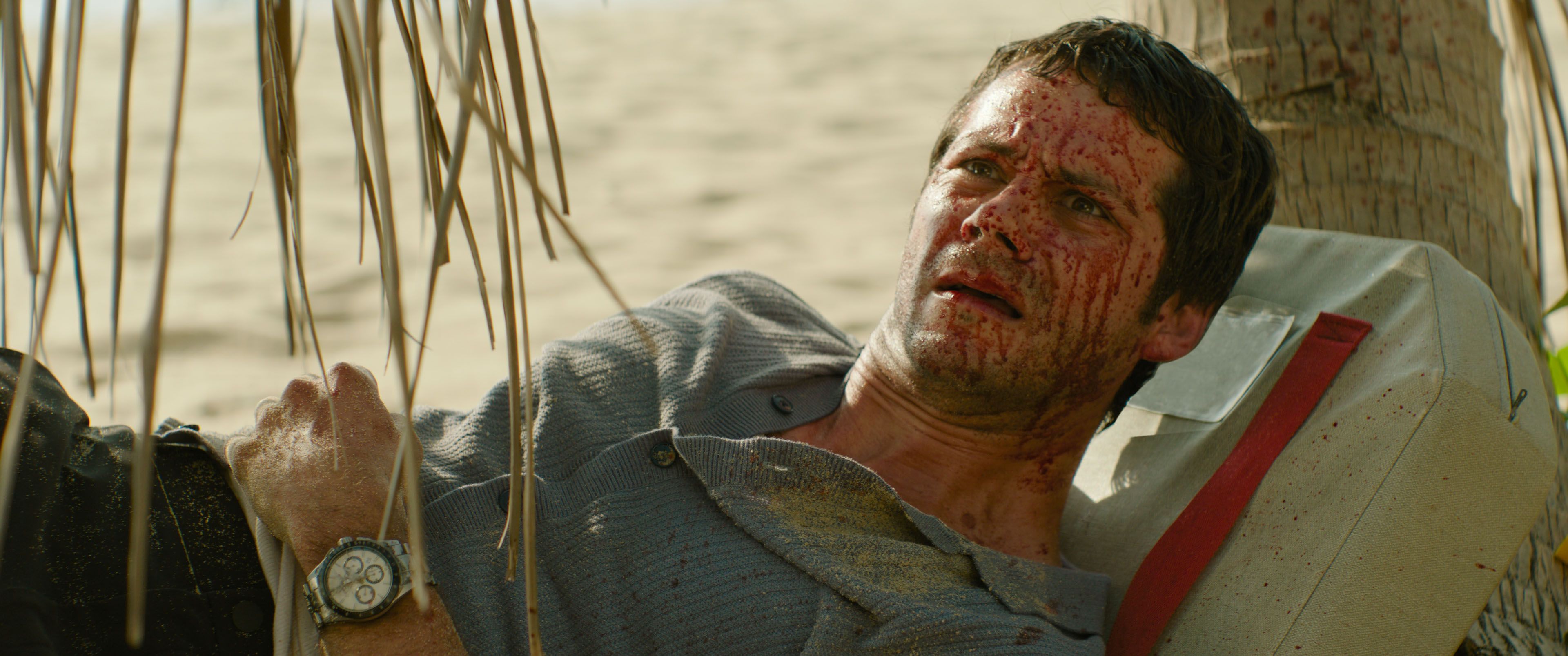 Bradley (Dylan O'Brien), a bedraggled man with a huge amount of blood spattered across his face, lies on a beach looking dumbstruck in Send Help