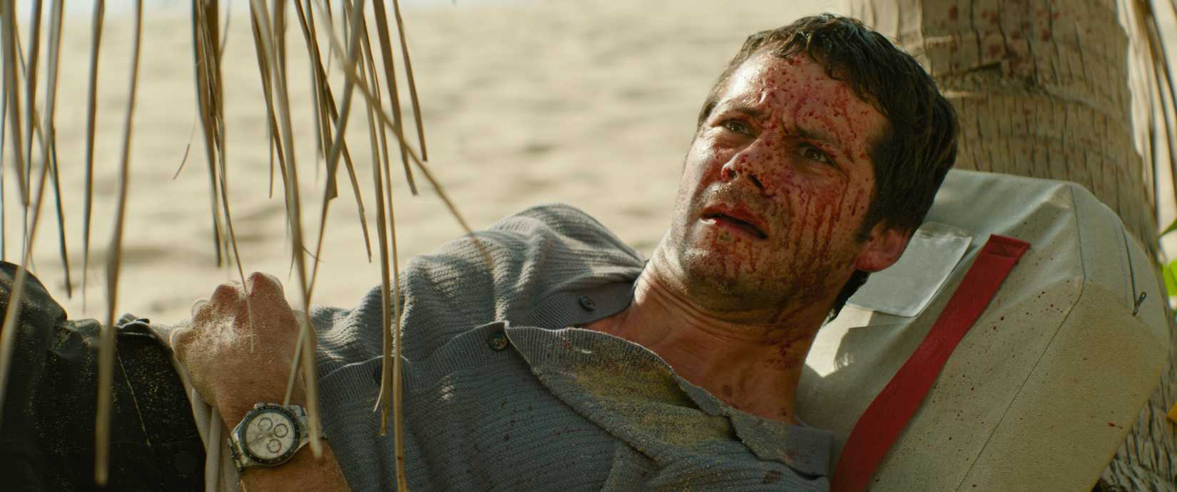 Bradley (Dylan O'Brien), a bedraggled man with a huge amount of blood spattered across his face, lies on a beach looking dumbstruck in Send Help