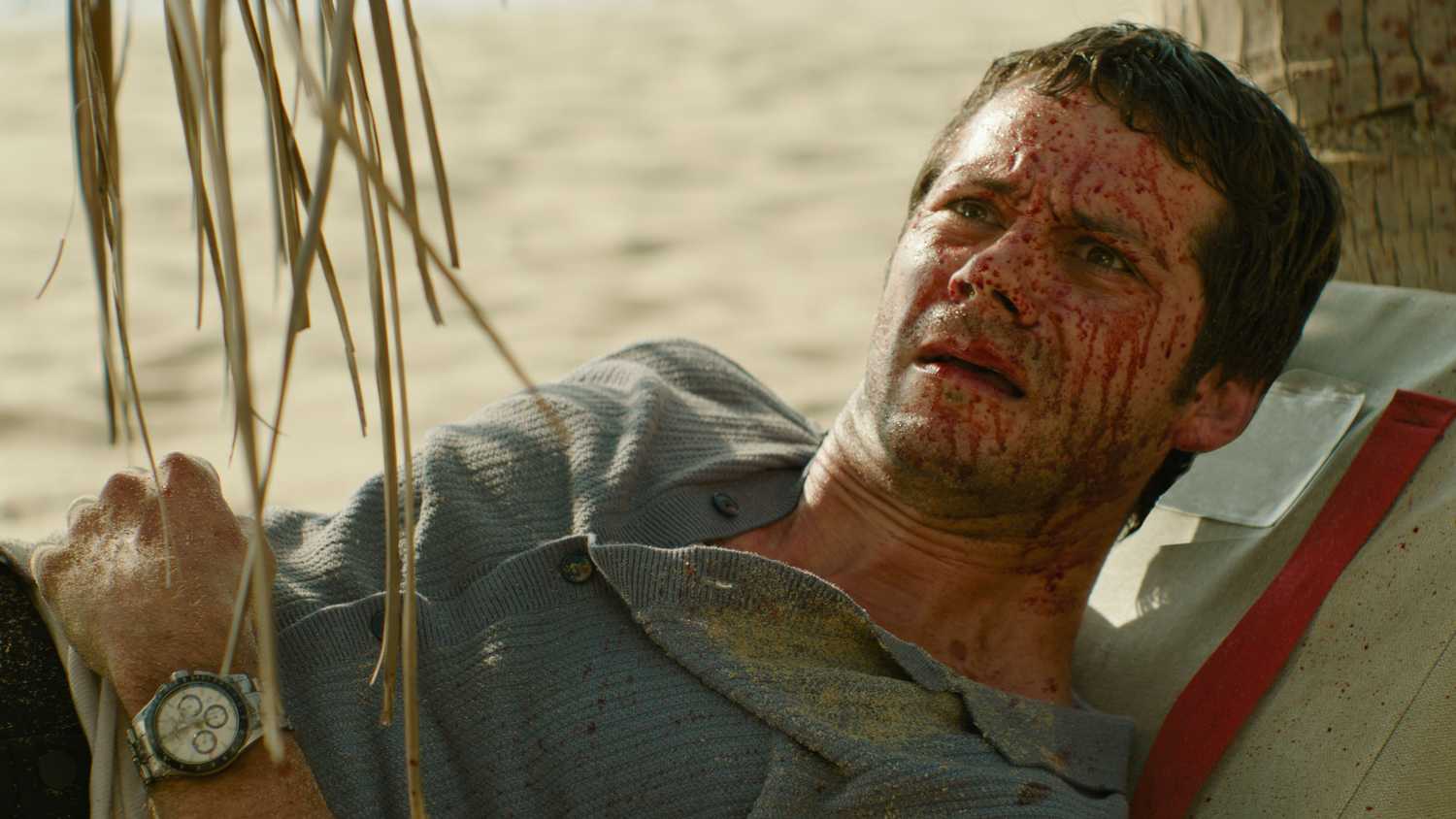Bradley (Dylan O'Brien), a bedraggled man with a huge amount of blood spattered across his face, lies on a beach looking dumbstruck in Send Help
