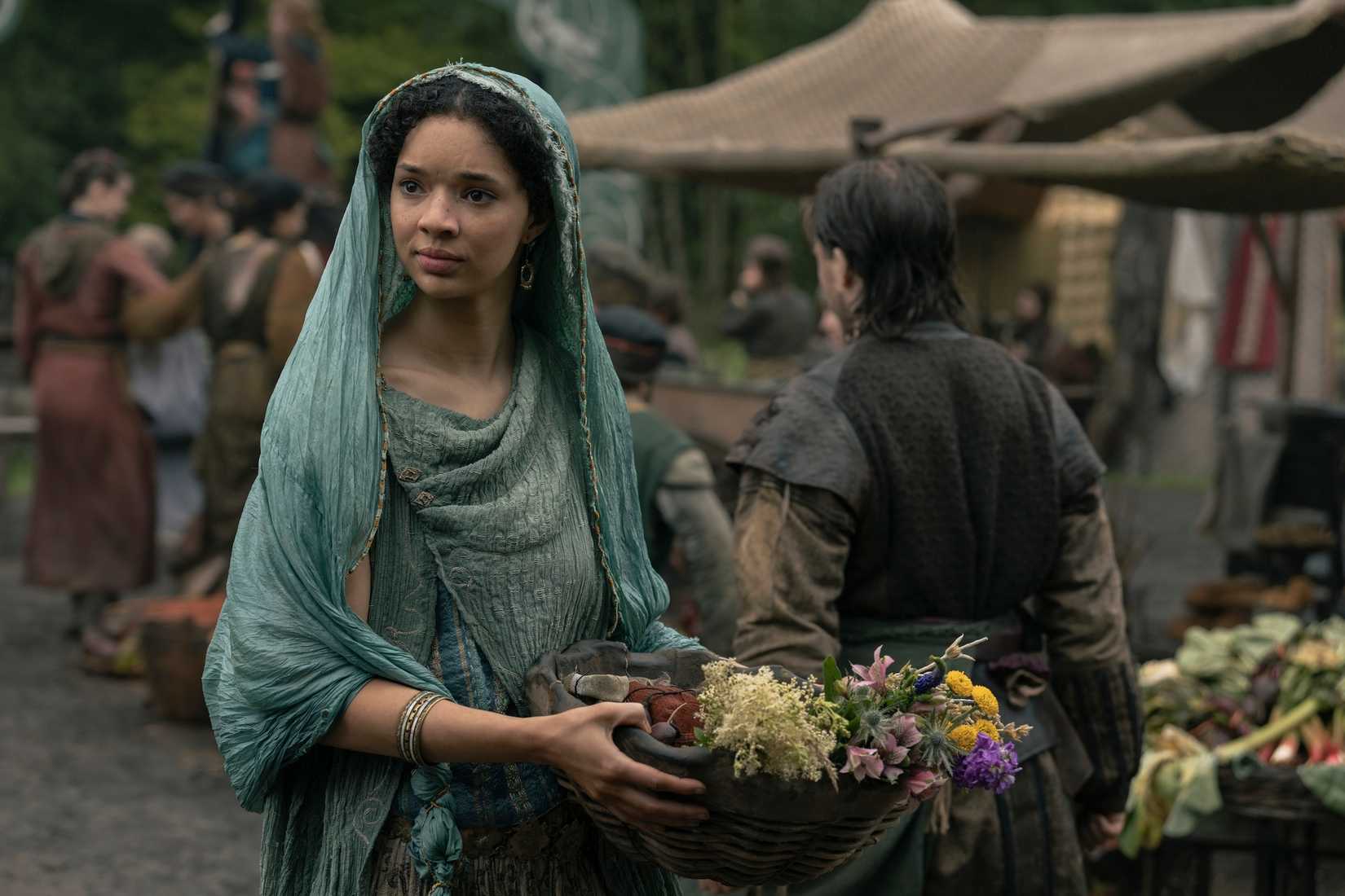 Tanselle (Tanzyn Crawford), a woman with long, dark hair, wearing a teal robe and headdress and carrying a basket of flowers, in A Knight of the Seven Kingdoms