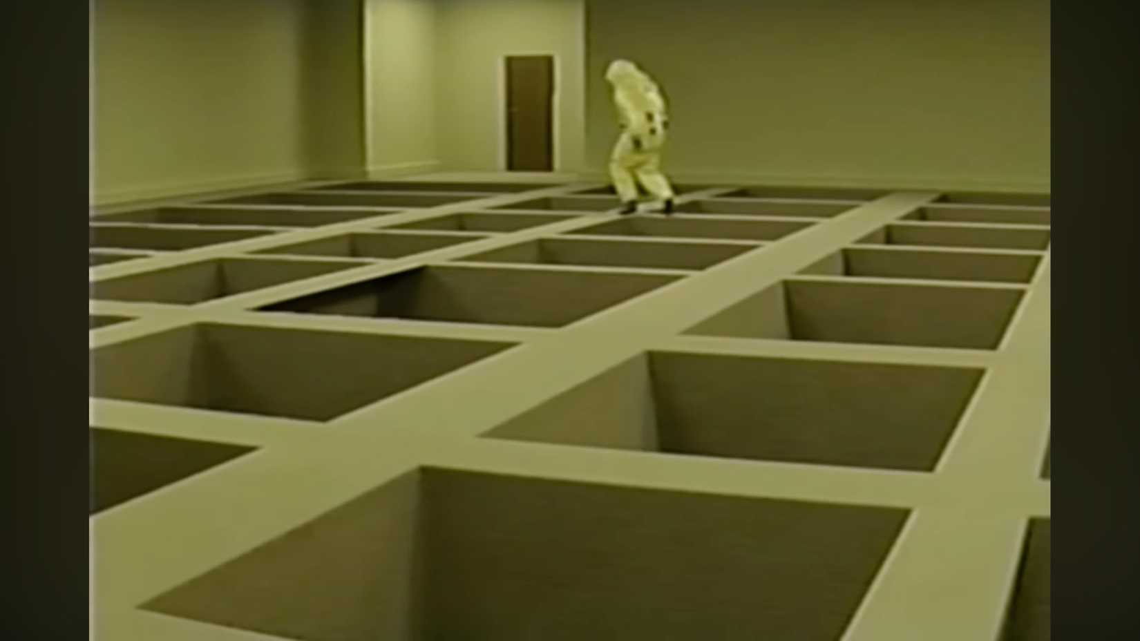 A researcher in protective gear observes massive square voids in the floor of a Backrooms level.