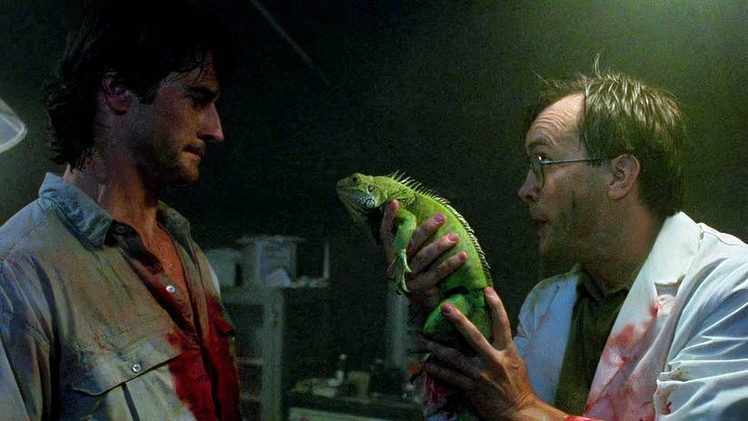 A bespectacled man (Jeffrey Combs) holds up an iguana to his friend (Bruce Abbott) in Bride of Re-Animator. Both men are liberally spattered with blood.