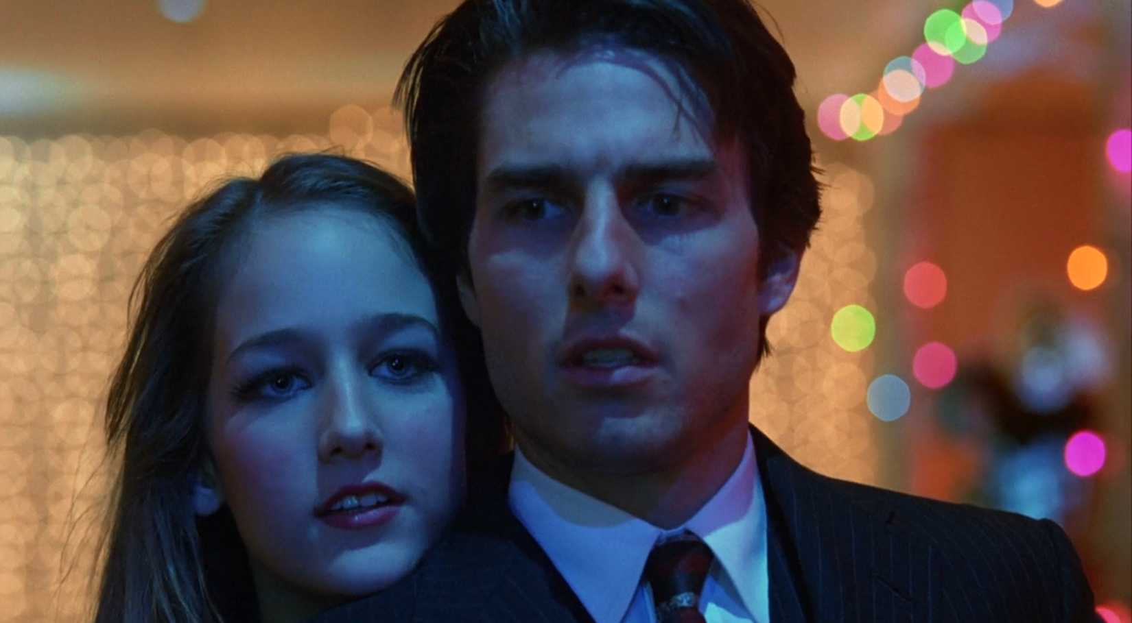 Bill (Tom Cruise) stands in a dimly lit room, wearing a suit and tie and looking unnerved. A young woman (Leelee Sobieski) hides behind him, her head on his shoulder, and stares off-screen. From Eyes Wide Shut.