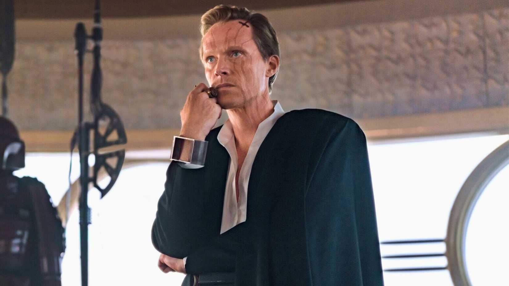  A Star Wars Story. Dryden is dressed in a black and white suit. He has a hand pressed to his face, and looks menacing.