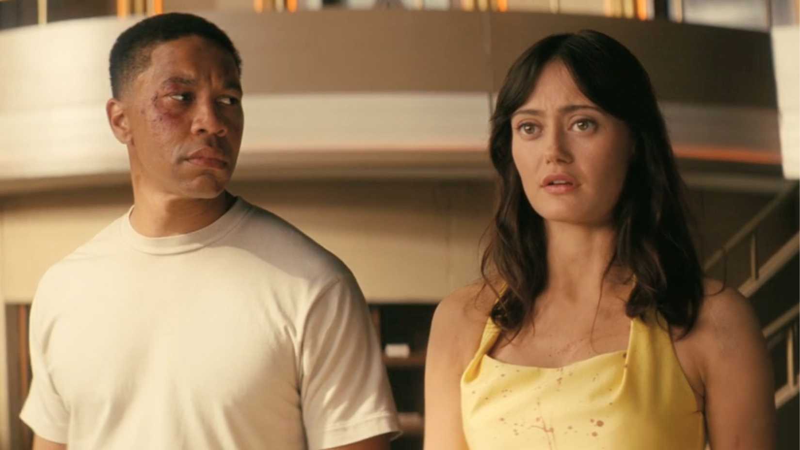 An image of Aaron Moten as Maximus and Ella Purnell as Lucy MacLean in Fallout season 2. Maximus, bruised and wearing a white shirt, looks at Lucy. She wears a yellow dress, covered with blood splatter.