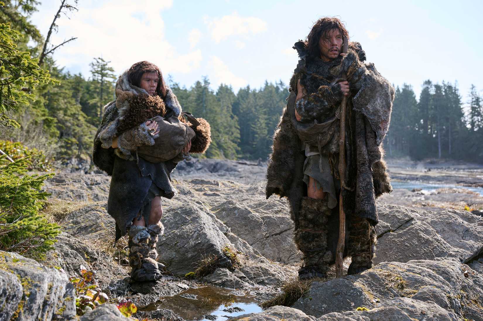 Actors playing Neanderthals, wearing clothing made of furs and skins, stand on a rocky beach in In the Blink of an Eye