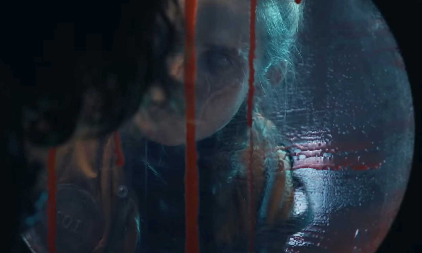 A woman with one completely white eye and a scar bisecting one cheek looks through a porthole dripping with blood in Iron Lung