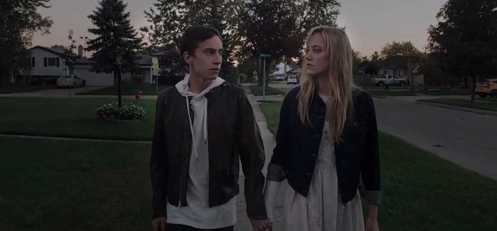 Paul and Jay, a young couple, hold hands and walk along the sidewalk in a suburban neighborhood, look at each other with blank faces and no sign of affection, in It Follows