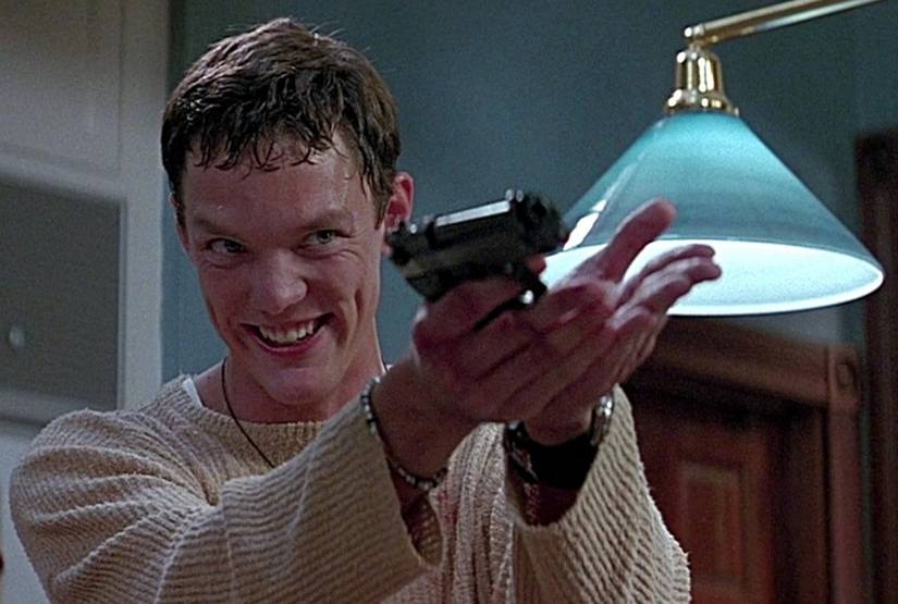 Stu Macher (Matthew Lillard), grinning and sweating, holds up a gun in a scene from the first Scream where he's revealed as one of the masked killers.