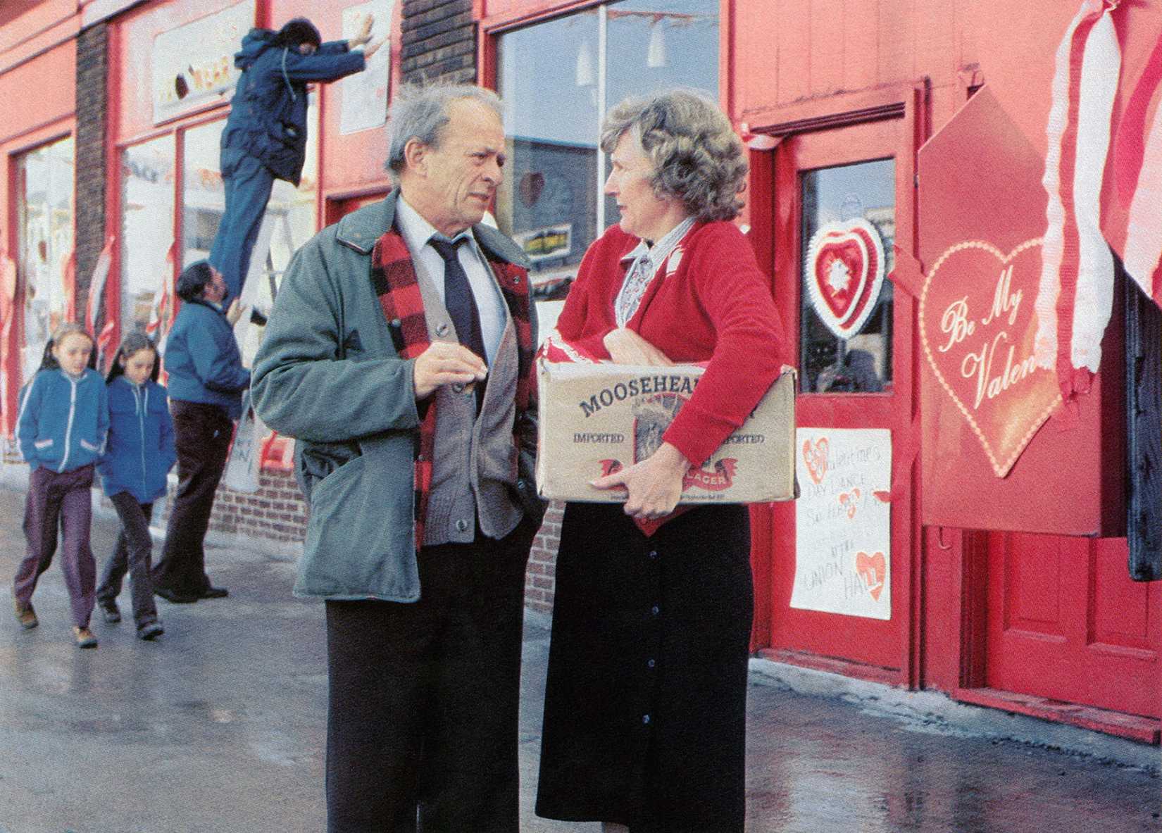 An image from the 1981 horror film, My Bloody Valentine. A man, dressed in black slacks, a grey jacket and cardigan, stands next to a woman. She wears a black skirt and red cardigan. They stand in front of a red store with valentine hearts stuck to it.