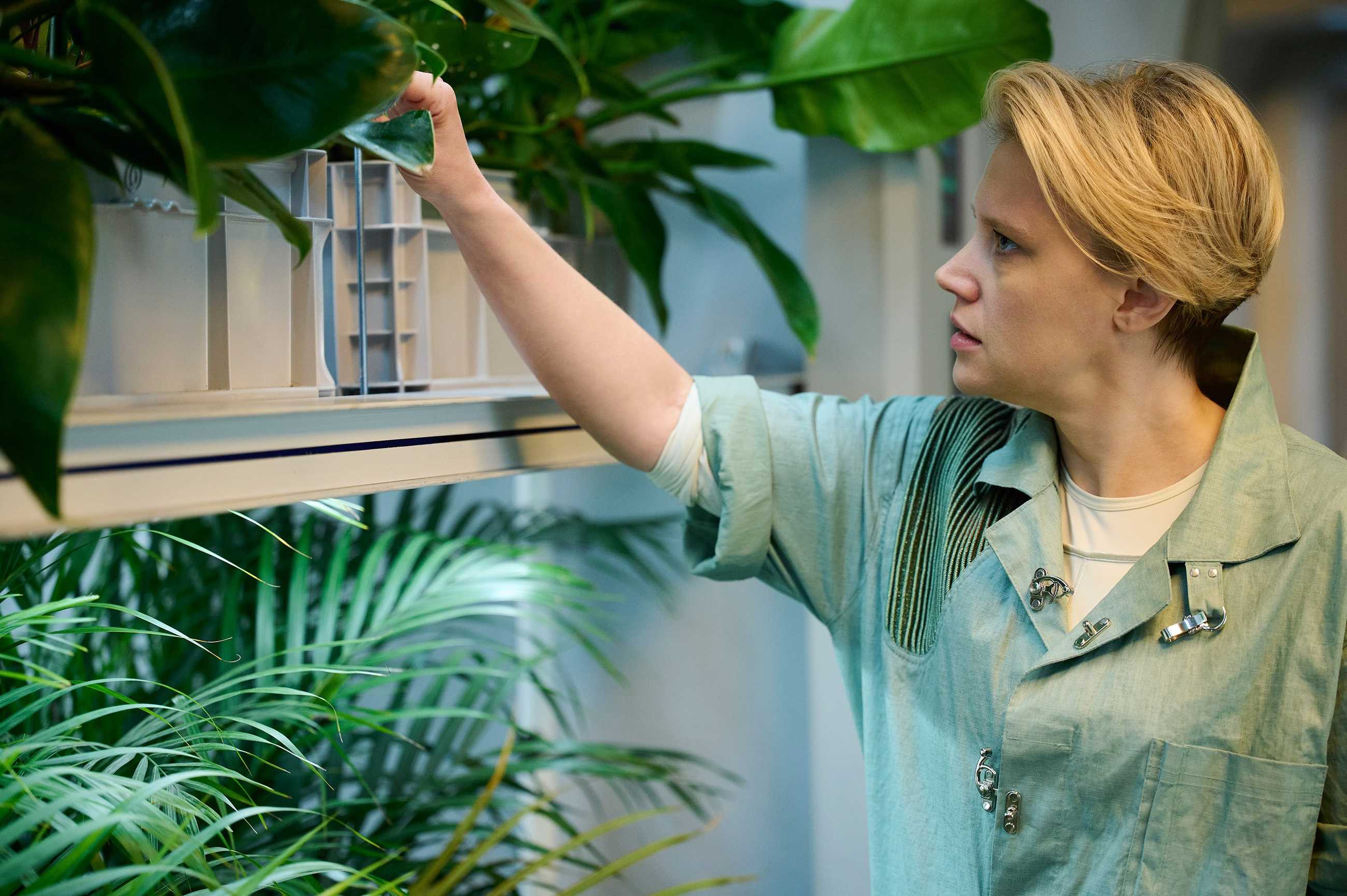 Coakley (Kate McKinnon), a woman with short blonde hair, wearing a soft, scrubs-like future jumpsuit, checks some plants with a worried expression in In the Blink of an Eye