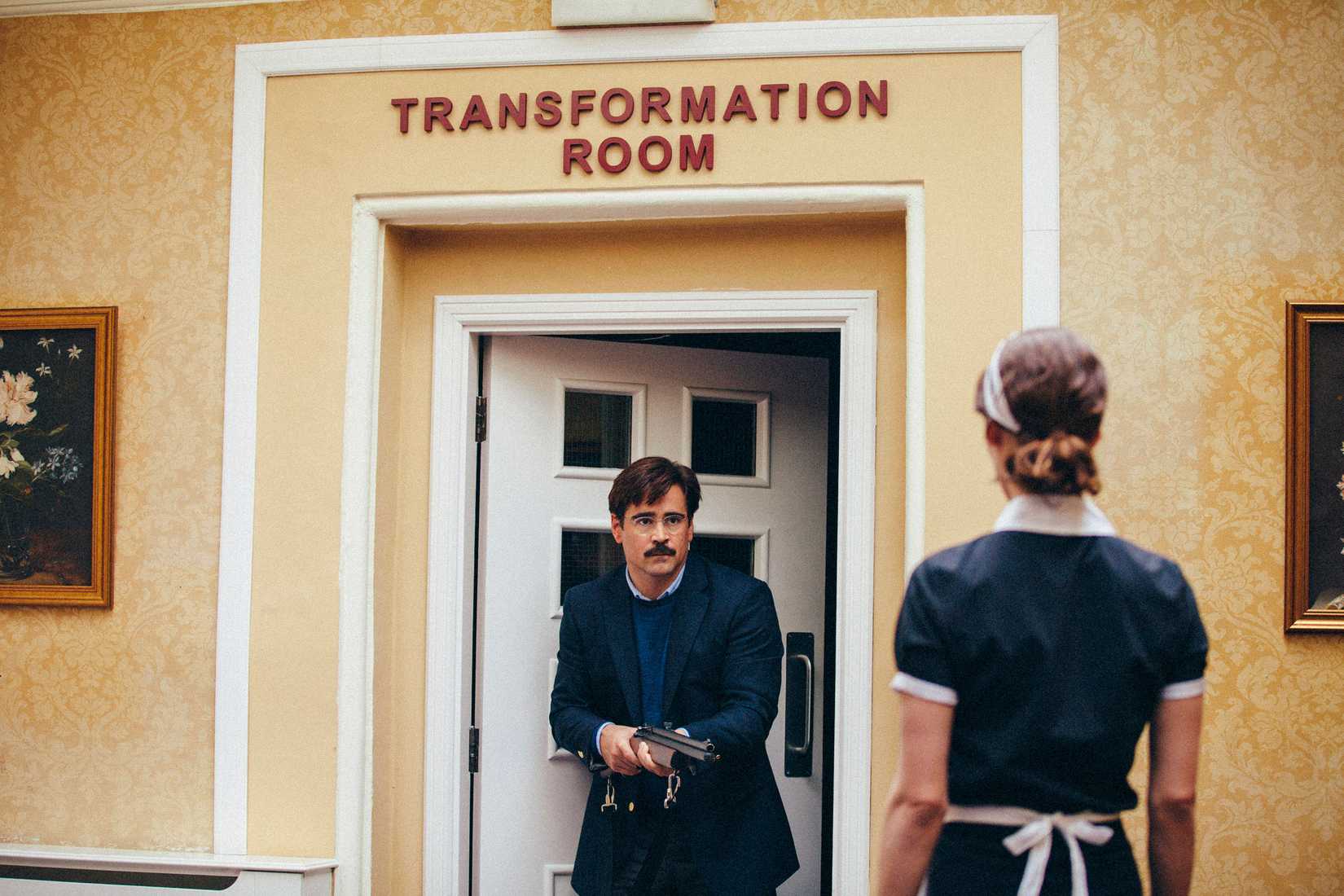 Colin Farrell holds a woman in a maid's outfit at bay with a shotgun while standing in a doorway below a sign that reads "TRANSFORMATION ROOM" in The Lobster