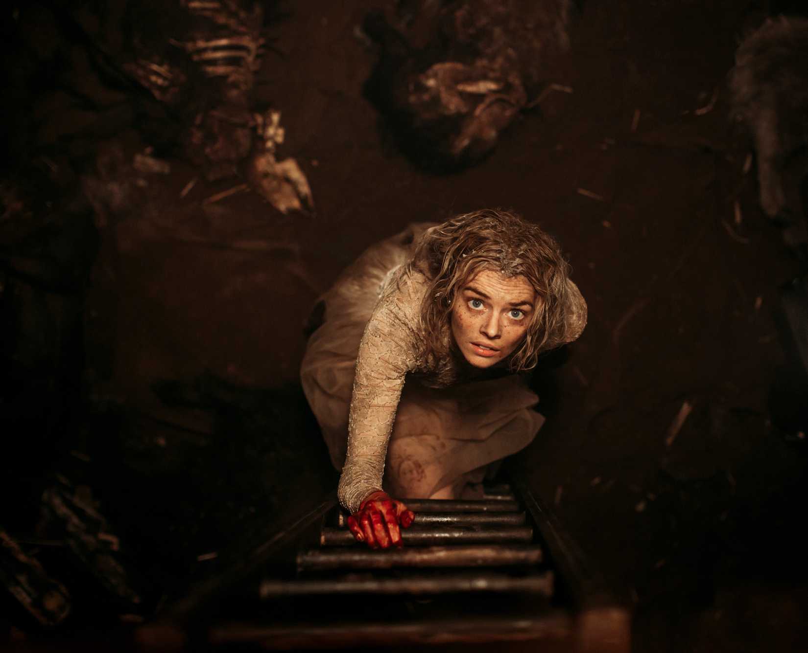 Samara Weaving, in a filthy white dress and with one hand covered in blood, starts up a grimy ladder from a dark, dirty space in Ready or Not