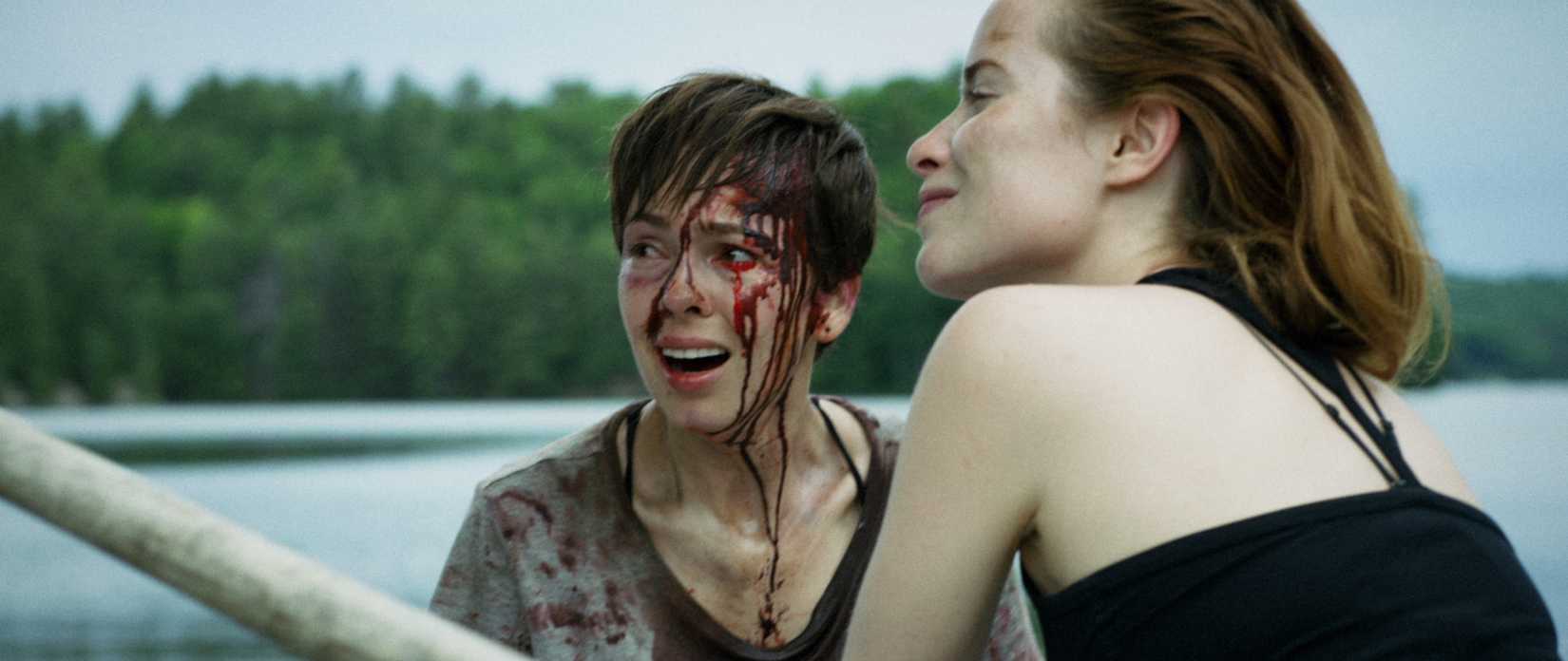 Brittany Allen, in a filthy, blood-covered shirt, with blood running down one side of her face and matted in her hair, smiles as Hannah Emily Anderson rows them in a boat on a lake in What Keeps You Alive.