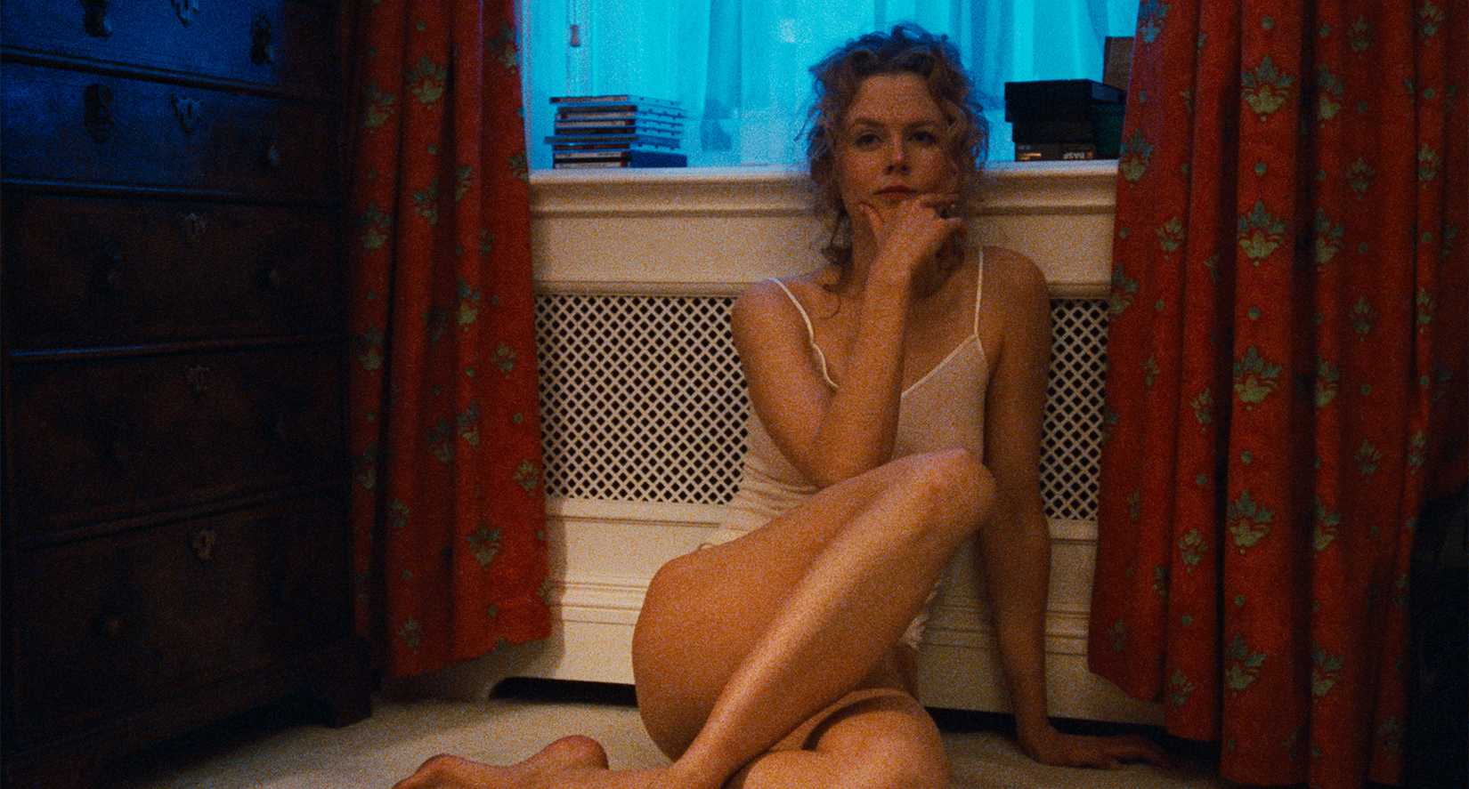 Alice (Nicole Kidman), wearing a thin white camisole and underwear, sits with her back to the wall, pensively holding her chin in her hand, in Eyes Wide Shut.
