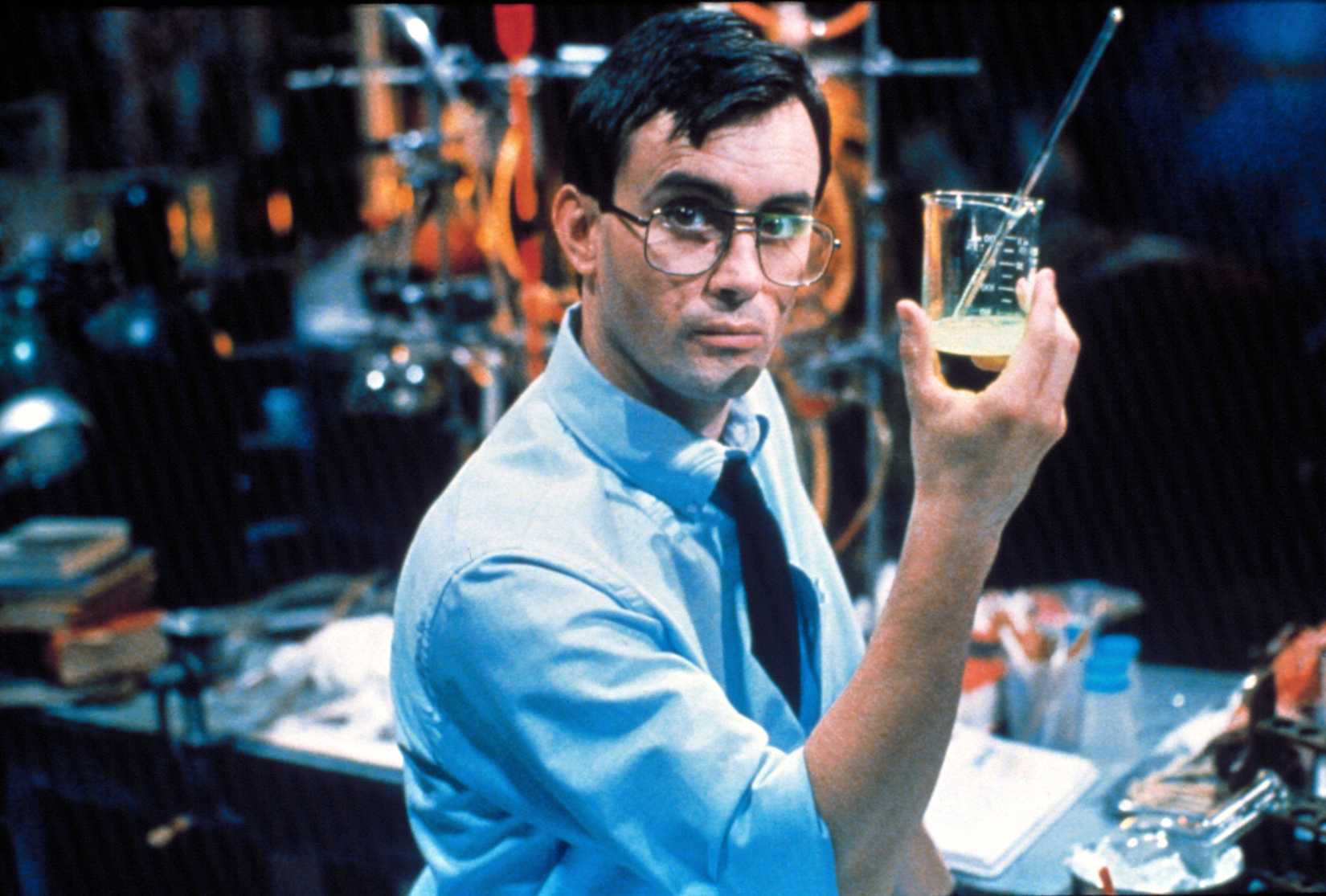 A bespectacled man (Jeffrey Combs) in a chemical lab looks toward the camera and holds up a beaker of liquid in Bride of Re-Animator