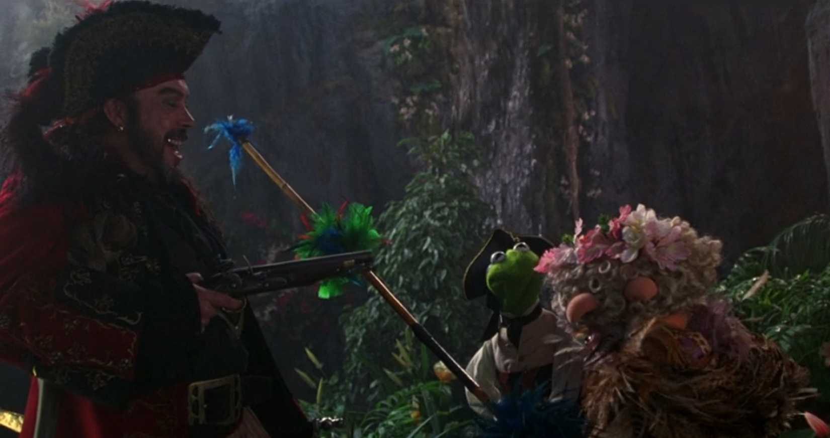 Long John Silver (Tim Curry) laughs as he brandishes a gun at Kermit and Piggy (who puts her head in her hands in distress/disgust) in a scene from Muppet Treasure Island