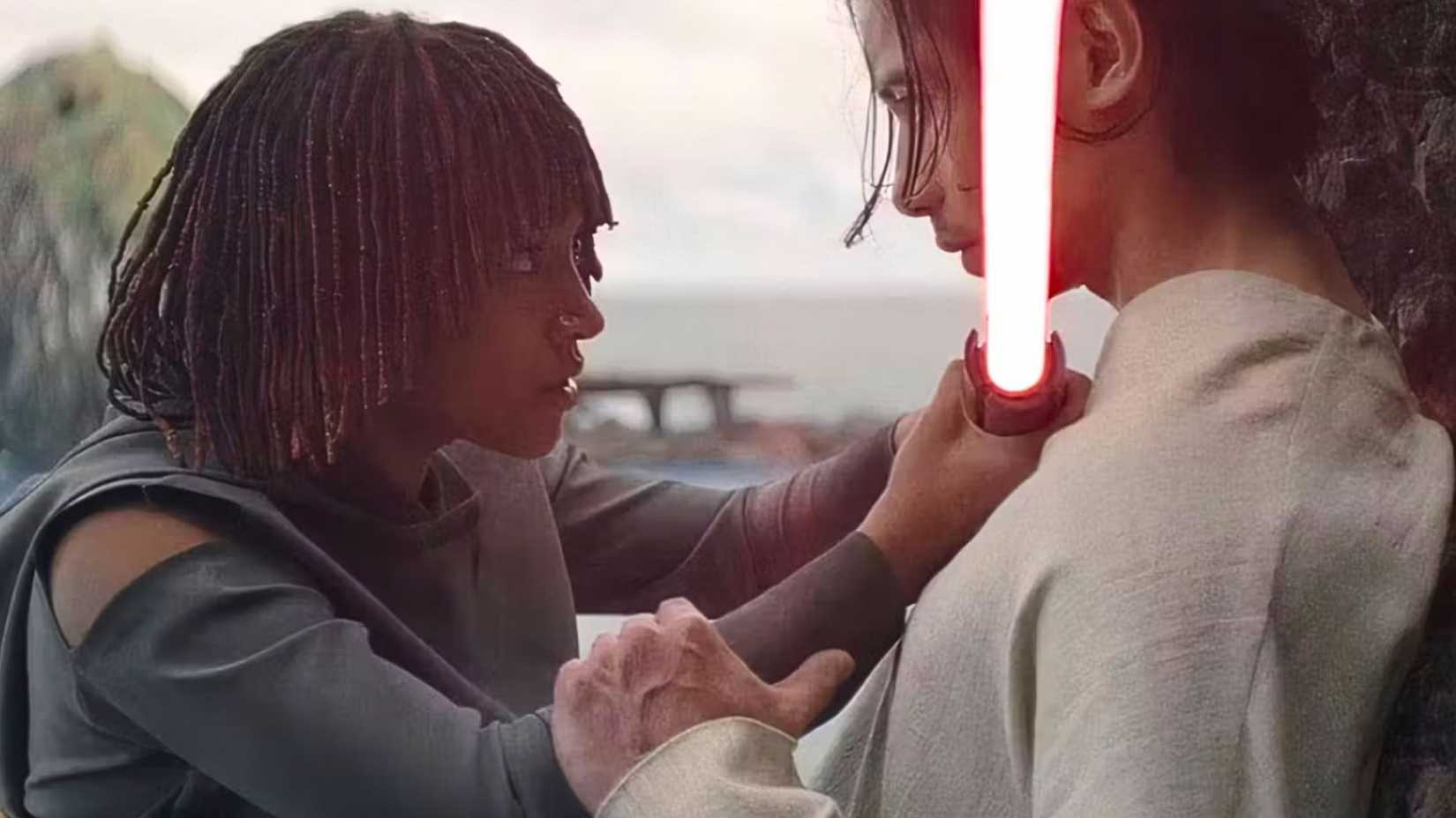 Osha and Qimir from Star Wars: The Acolyte. Osha, wearing a black/grey outfit, holds up a red lightsaber to Qimir's throat. Qimir, wearing white, holds onto her wrist. 