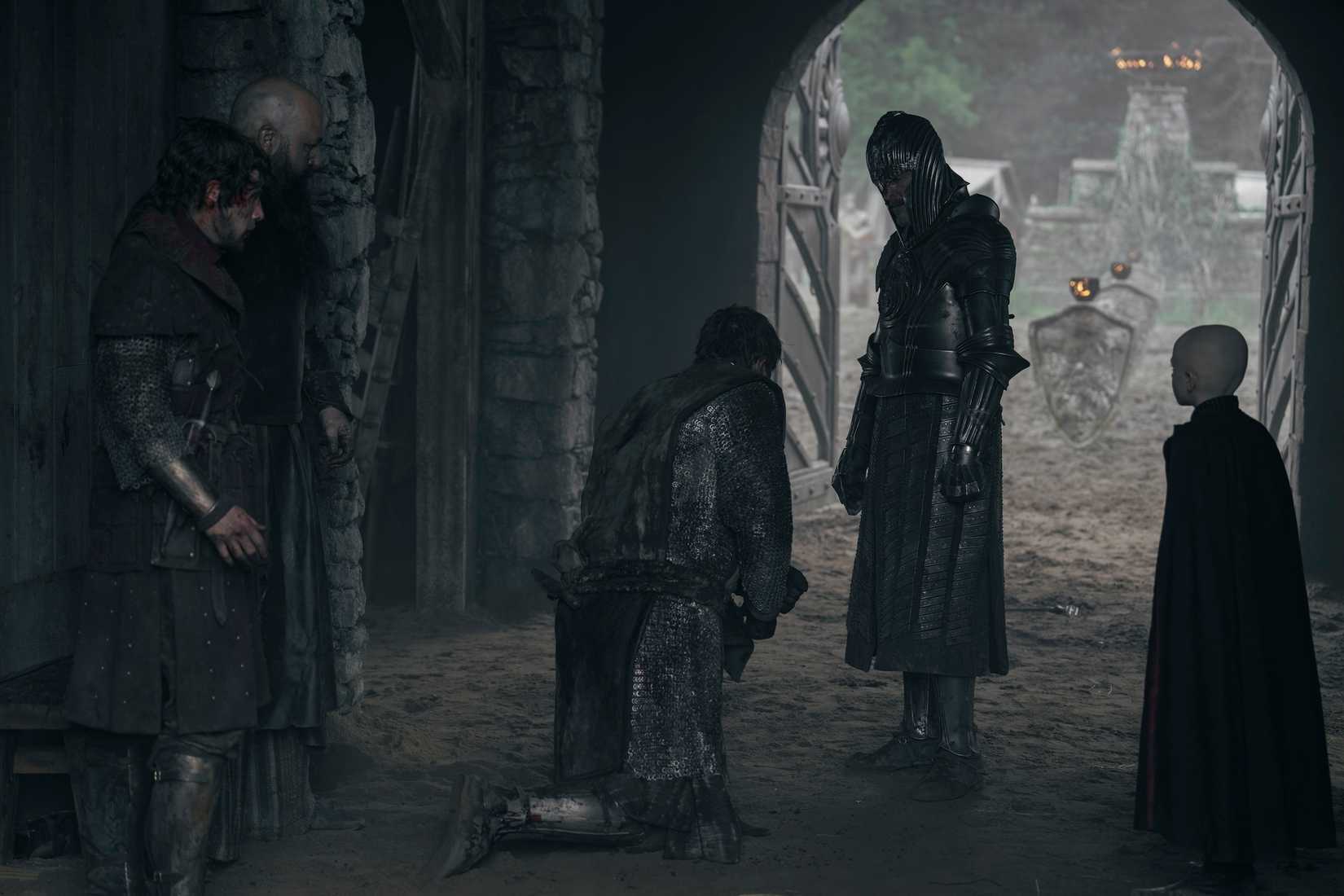 A group of men, several in medieval plate armor, and a small bald boy stand in a dark, shadowy stable, with one man kneeling in the middle, in A Knight of the Seven Kingdoms