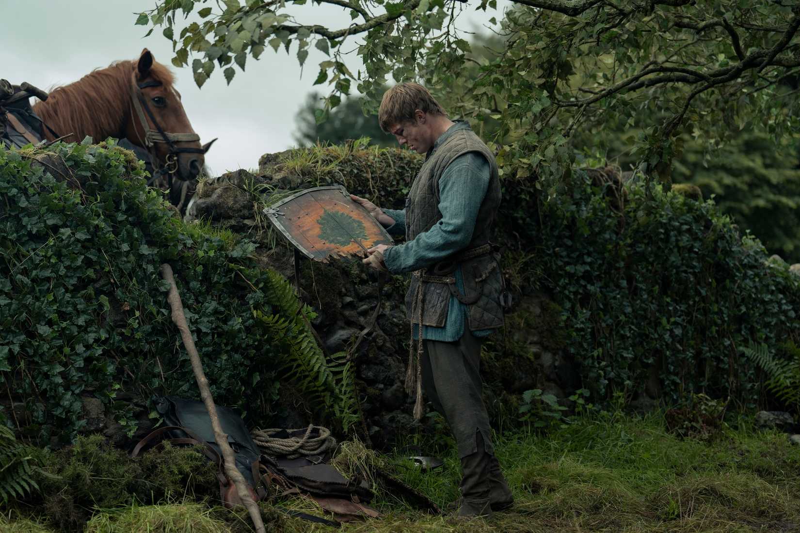 Dunk (Peter Claffey), a tall, battered man in medieval clothing, holds a broken wooden shield up and examines it. He's standing in front of a hedge with a horse peeking over it. From A Knight of the Seven Kingdoms.