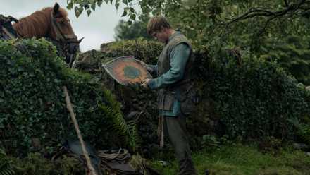 Dunk (Peter Claffey), a tall, battered man in medieval clothing, holds a broken wooden shield up and examines it. He's standing in front of a hedge with a horse peeking over it. From A Knight of the Seven Kingdoms.