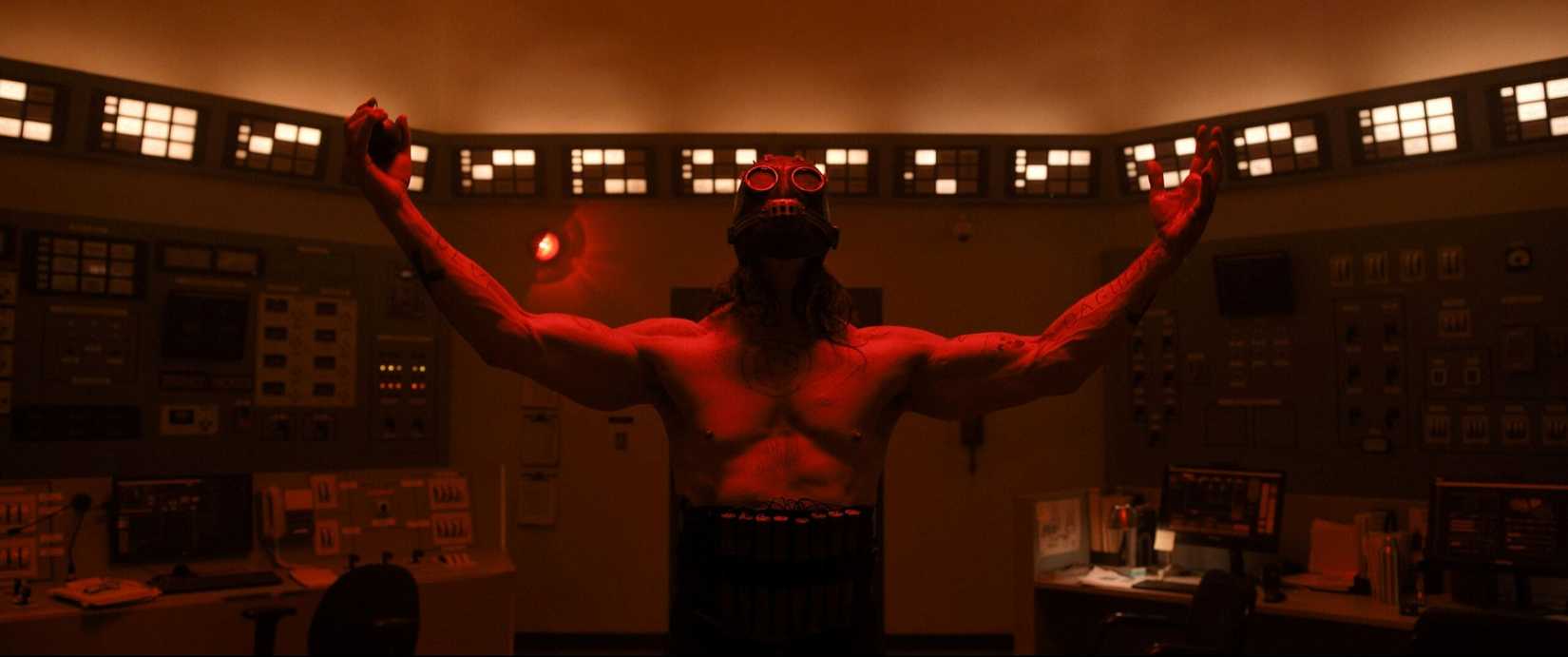 A gas-masked, shirtless killer stands with his arms outstretched in a room bathed in red light in a scene from the movie Psycho Killer.