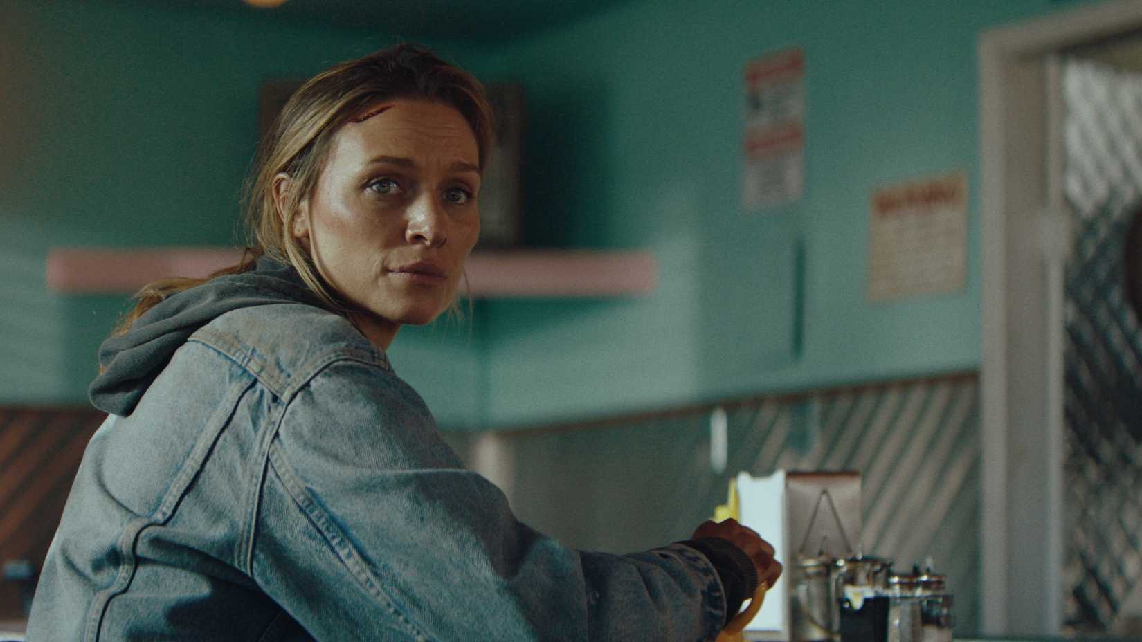 Irene (Michaela McManus), a middle-aged woman wearing a jean jacket with a cut on her head, looks over her shoulder while sitting in a diner in Redux Redux.