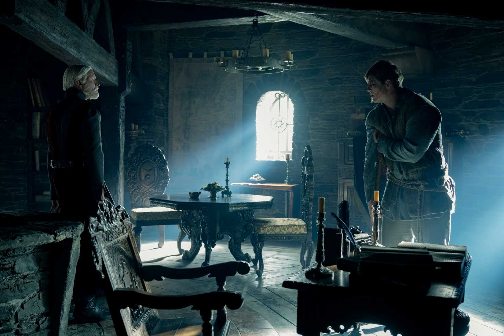 Two men, one older and dressed in black, one younger, badly bruised, and leaning on a crutch, face either other in a very dark medieval-ish room with light pouring in through a central window in A Knight of the Seven Kingdoms