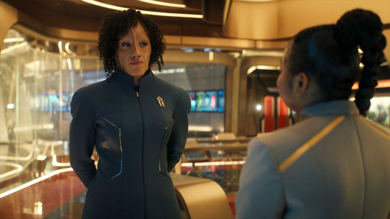 Illa Dax (Tawny Newsome) a Cardassian in a blue uniform, stands in a classroom talking to Sam (Kerrice Brooks) a student in a grey uniform