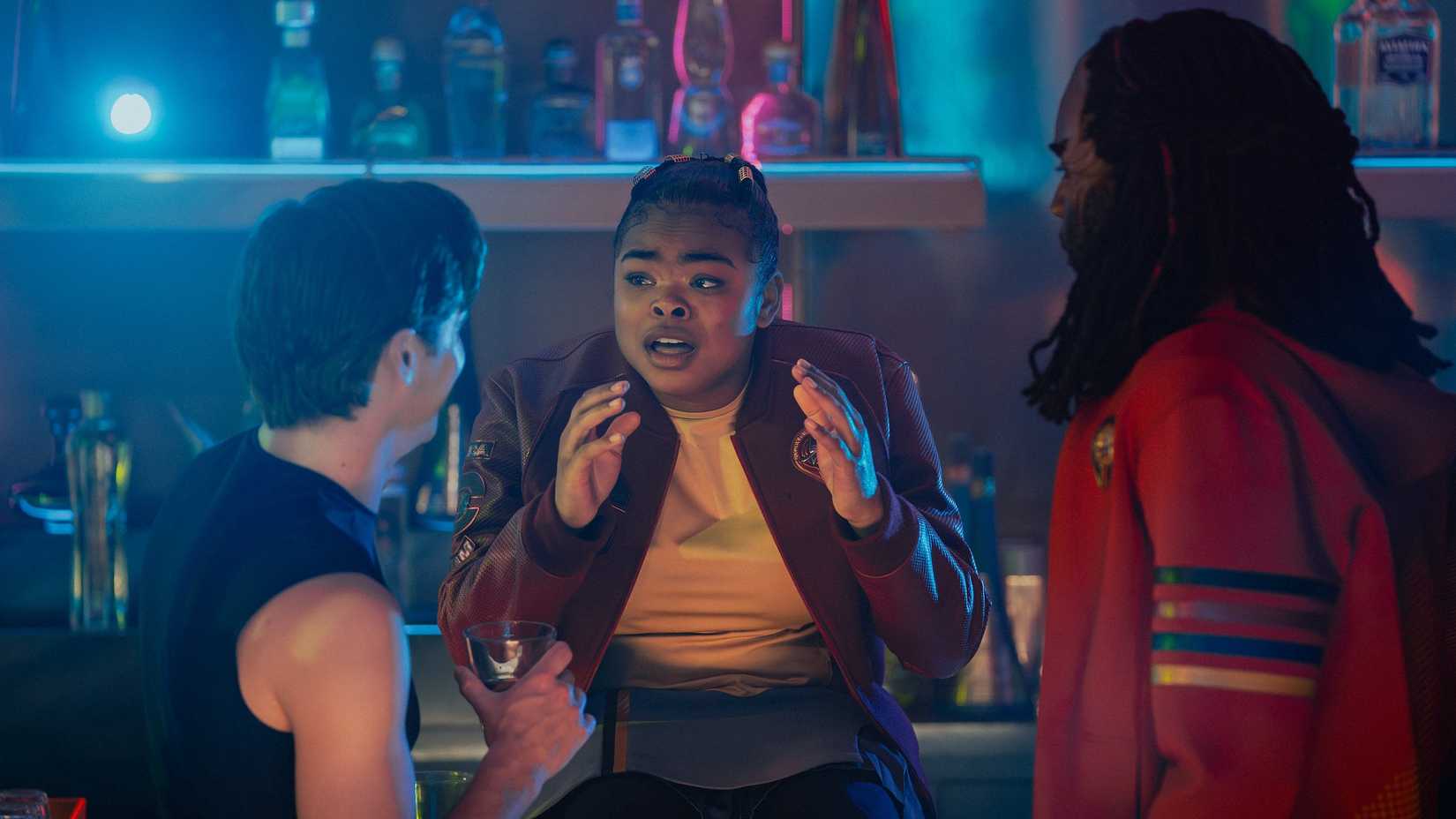 Sam (Kerrice Brooks) a young drunk woman dressed in a skirt and tights sits on a bar and talks to two fellow students