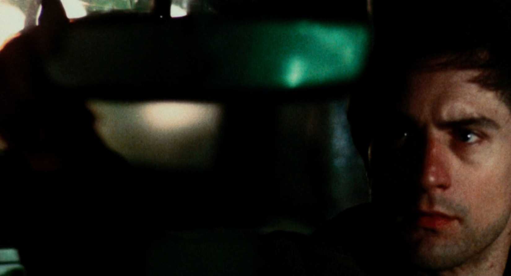 The face of Travis Bickle (Robert De Niro) is visible in the bottom right corner of an otherwise distorted, blurry view of his taxicab and the outside world, as he looks in the rearview mirror at something the audience can't see.