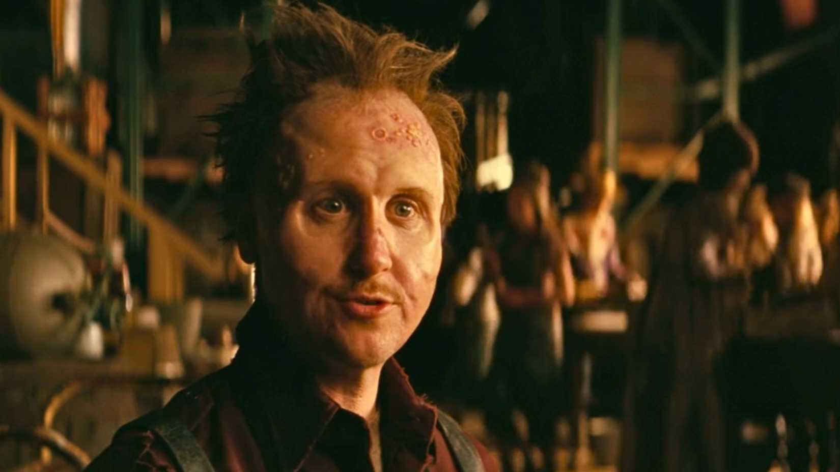 An image of Thaddeus from Fallout season 2. He has short hair that sticks up and a face that looks scarred and pockmarked. 