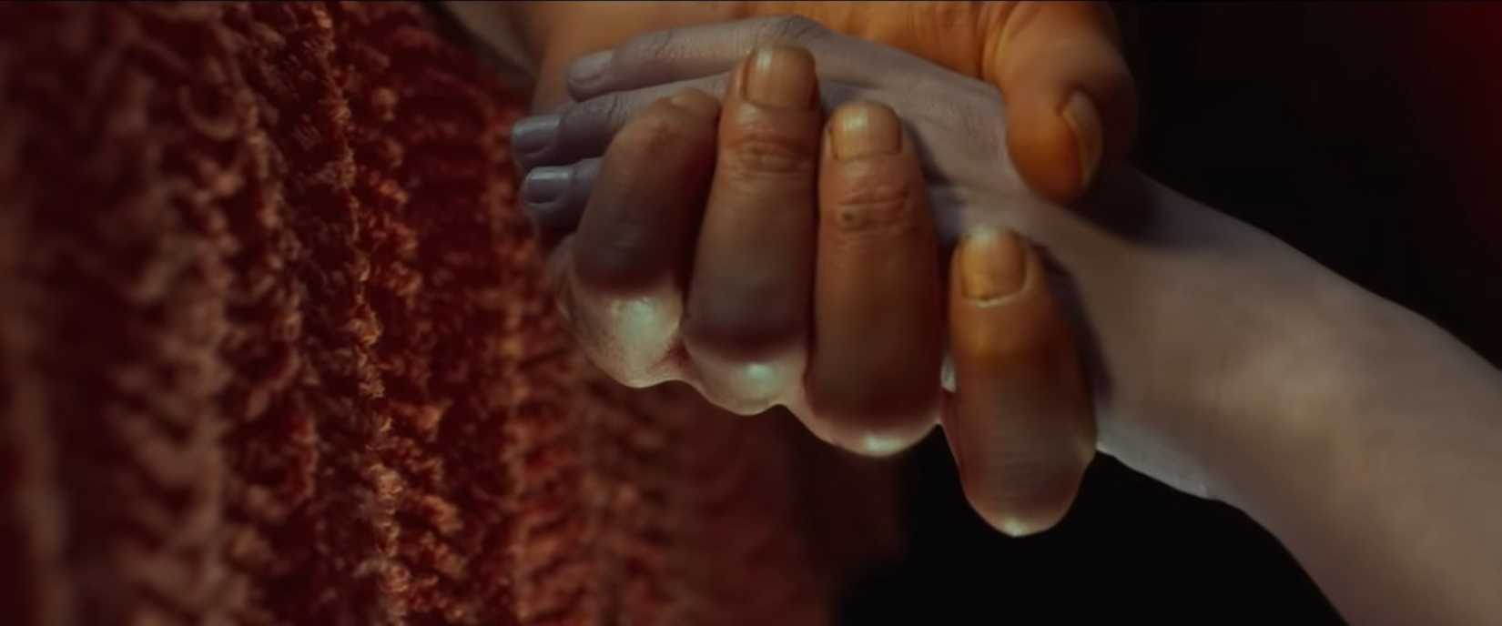 Two hands, an adult hand that looks orange-y in color and a child's hand that's more pale and grey, hold each other in close-up in a scene from Alpha.