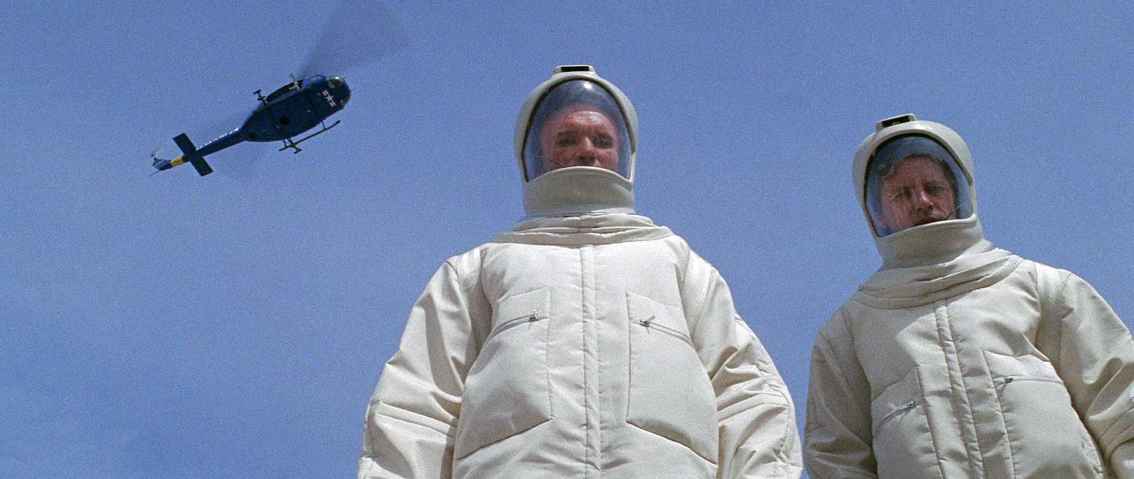 Two men in Hazmat suits stare at the ground as a helicopter passes in The Andromeda Strain