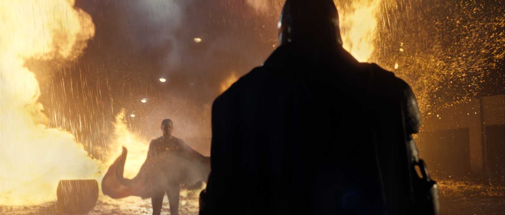 Superman standing far away amidst rain and fire while Batman stands in the foreground with his back, and Bat Cape, turned toward the camera in an image from Batman v Superman Dawn of Justice.