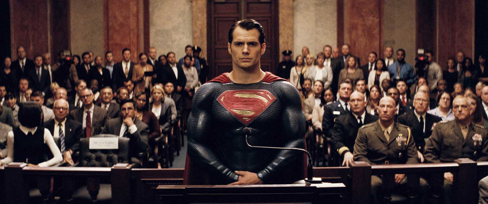 A very moody Superman standing with his hands clasped at his waist inside the Capitol Building, with dozens of seated audience members behind him, in an image from Batman v Superman Dawn of Justice.