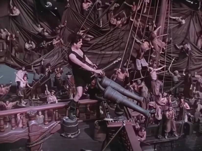 A wide shot of a man (Douglas Fairbanks) disguised as a pirate, standing on the upper deck of a ship, aiming a pair of cannons at the many sailors below him.