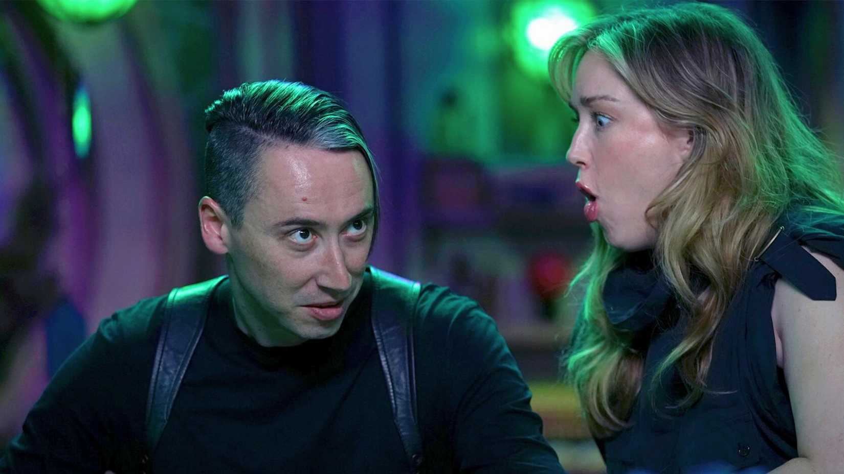 An image from Critical Role showcasing Alex Ward and Ashley Johnson. Alex has a serious look on his face, dressed all in black. Ashley, next to him, pulls a face of shock. 