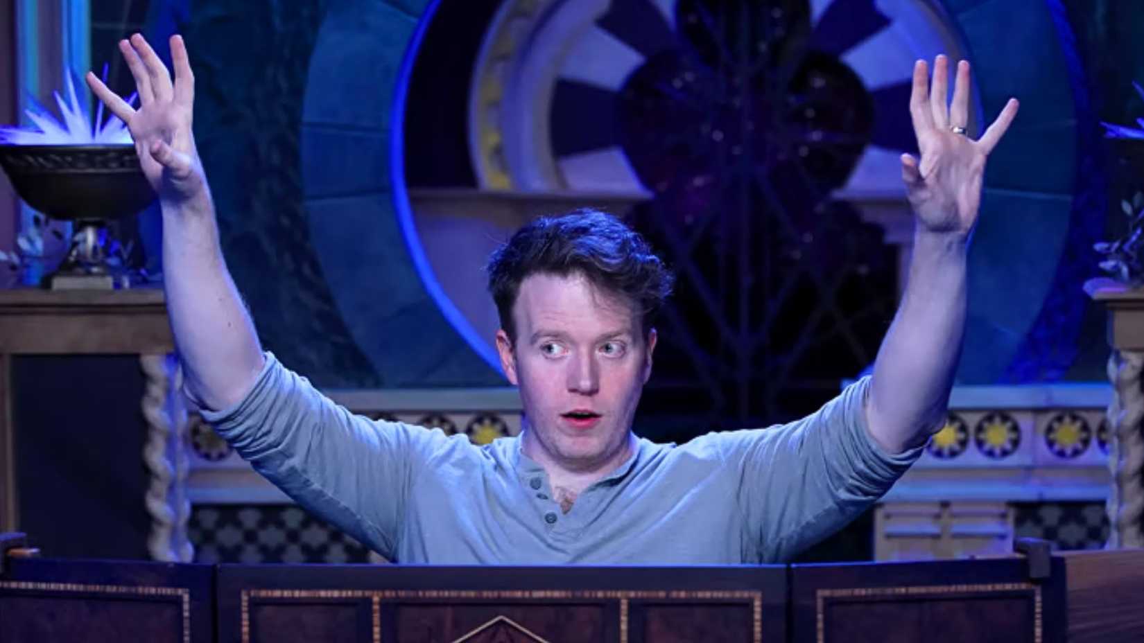 An image featuring Brennan Lee Mulligan behind a Dungeon Master screen. Behind him is a fantasy background, awash with blue light.