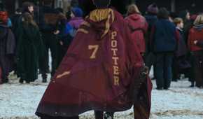 Harry Potter (Dominic McLaughlin), seen from behind as he walks across a snowy courtyard into Hogwarts, with a crowd of other out-of-focus students in the background. He's wearing a cloak that says "7" and "Potter" in the Gryffindor House colors.