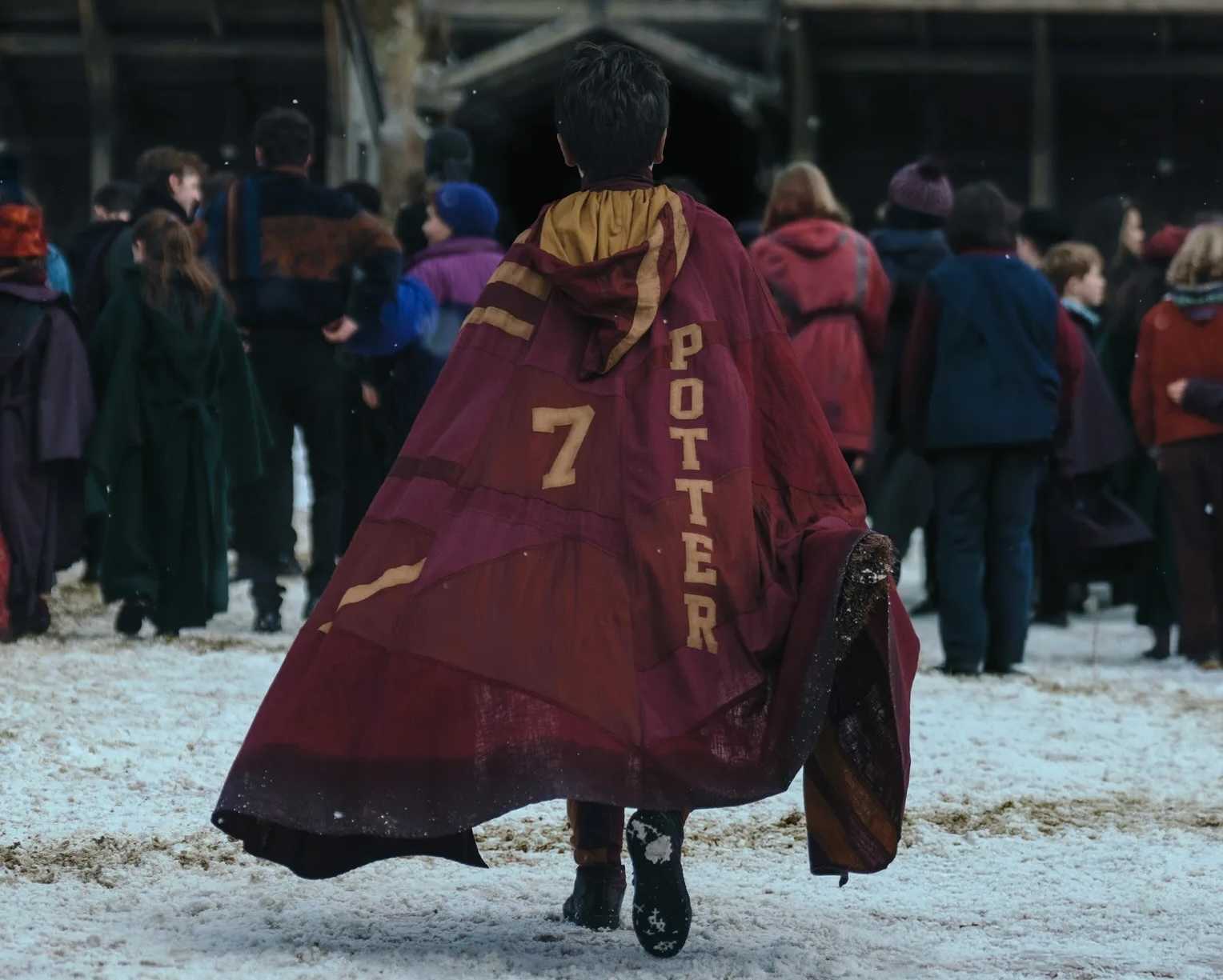 Harry Potter (Dominic McLaughlin), seen from behind as he walks across a snowy courtyard into Hogwarts, with a crowd of other out-of-focus students in the background. He's wearing a cloak that says "7" and "Potter" in the Gryffindor House colors.