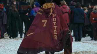 Harry Potter (Dominic McLaughlin), seen from behind as he walks across a snowy courtyard into Hogwarts, with a crowd of other out-of-focus students in the background. He's wearing a cloak that says "7" and "Potter" in the Gryffindor House colors.