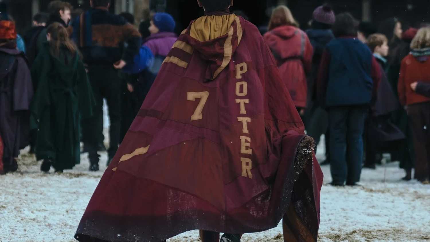 Harry Potter (Dominic McLaughlin), seen from behind as he walks across a snowy courtyard into Hogwarts, with a crowd of other out-of-focus students in the background. He's wearing a cloak that says "7" and "Potter" in the Gryffindor House colors.