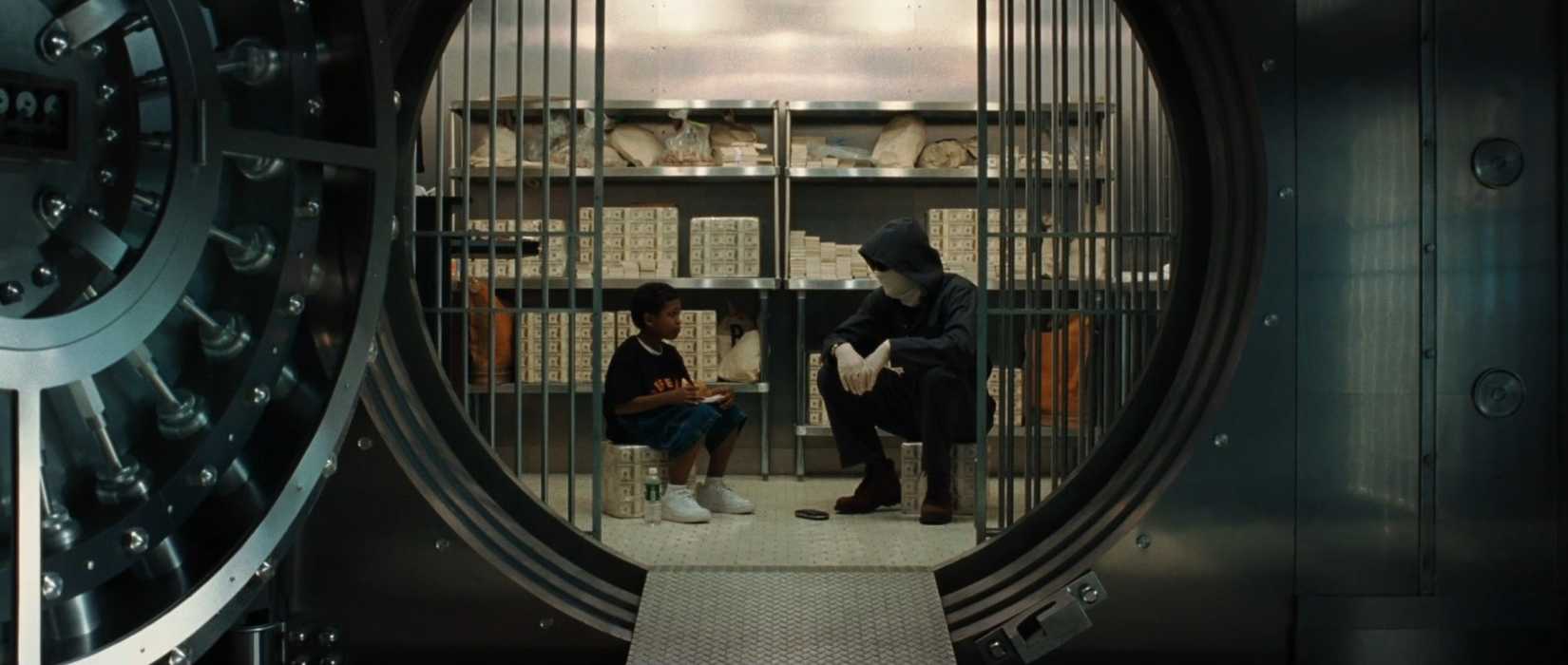 In a scene from the movie Inside Man, shot from a distance, a bank robber played by Clive Owen sits in a bank vault and chats with a tweenage boy. They face each other center of frame, with the circular opening to the value surrounding them and further framing the image.