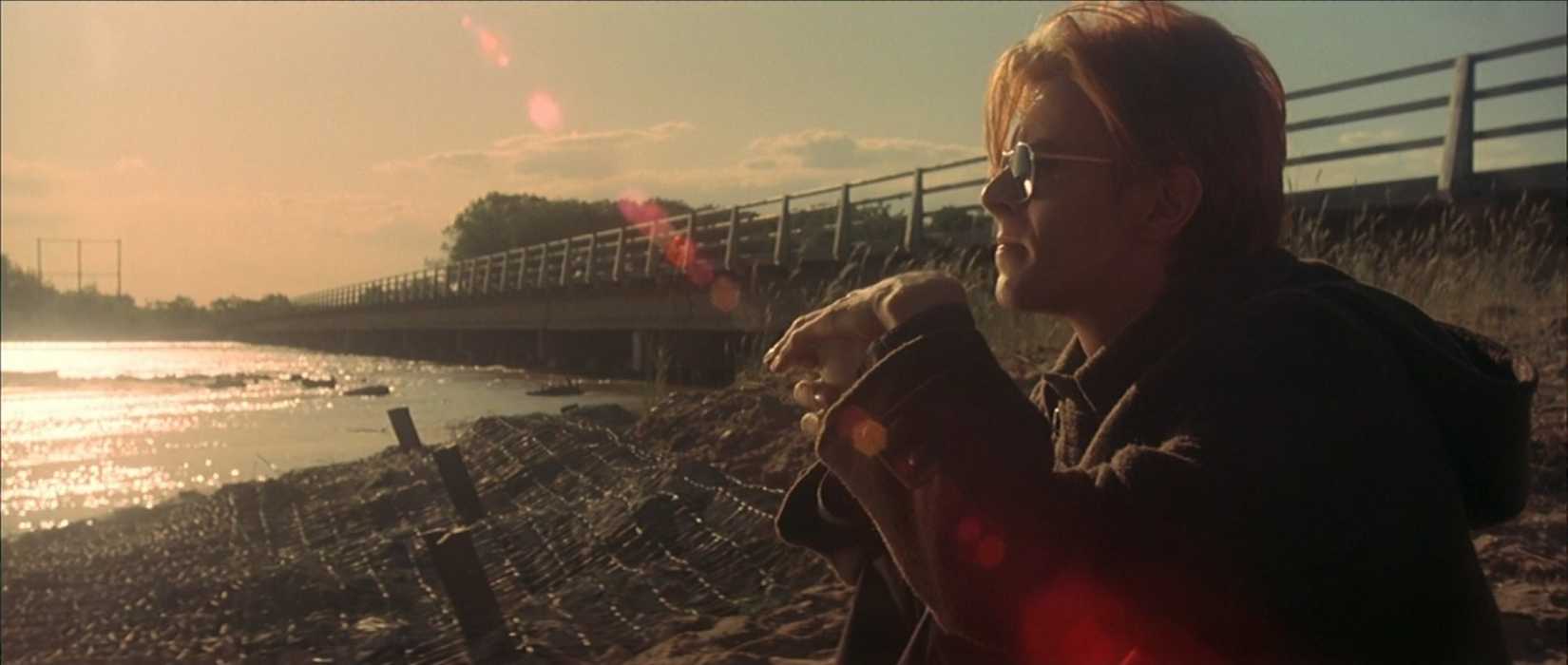 An alien in human form (played by David Bowie, of course) sits on a lake shore, wearing sunglasses and looking contemplative as the sun shine behinds him in a scene from The Man Who Fell to Earth (1976).