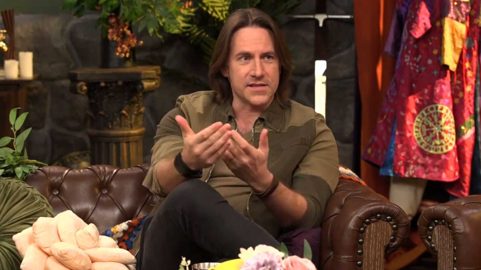 An image showing Matthew Mercer sitting on a couch. He wears a green and brown jacket and his hair just falls above his shoulders. He moves his hands as he explains something.