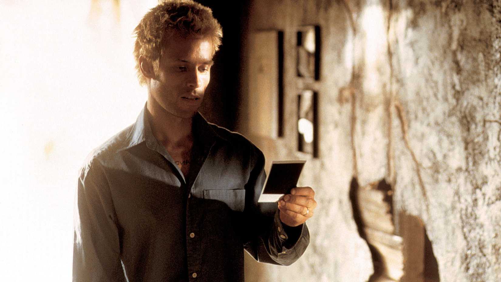 Leonard Shelby (Guy Peace), stands in a worn down building holding a Polaroid photo in Memento.